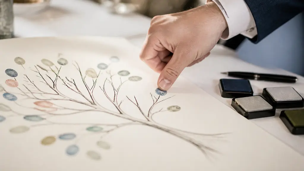 Close-up editorial photograph of a wedding guest pressing an inked thumb onto a paper tree illustration, colorful fingerprint leaves on the branches, black pen and ink pads beside the flat artwork