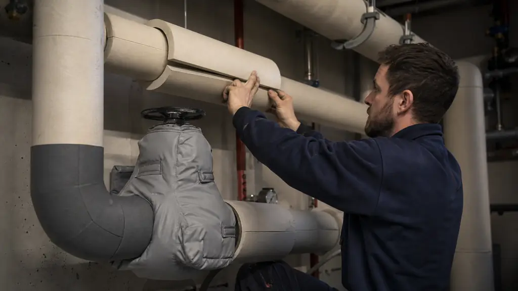 Editorial photograph of a technician fitting rigid insulation shells onto a straight heating pipe beside a removable insulated valve mattress and a carefully shaped foam-covered elbow, no text or logo