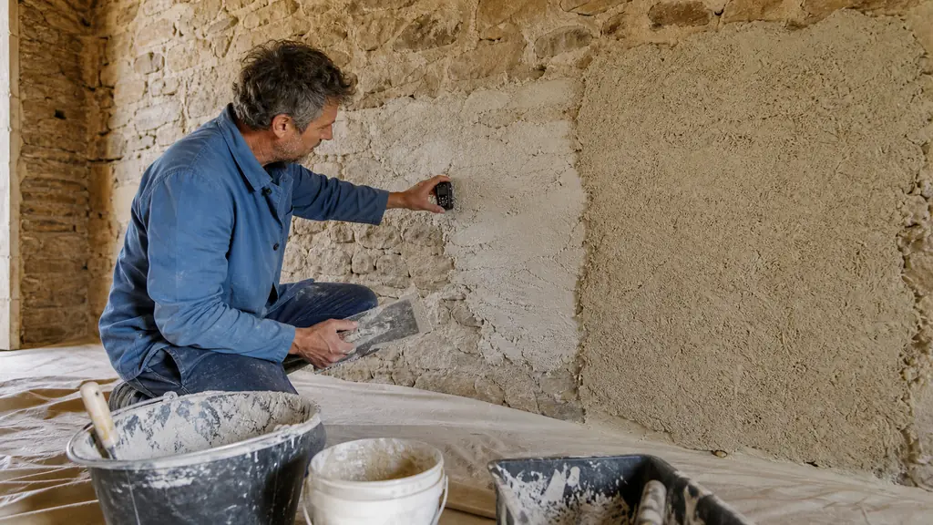 editorial photograph of an artisan checking moisture on an exposed old stone wall while applying a rough gobetis coat beside a fresh lime-hemp plaster test area, no text, no logo